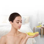 A woman holding a discreet banana-shaped clitoral stimulator in a bright, modern bedroom setting, showcasing the product’s playful aesthetic and lifestyle integration for personal wellness.