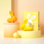 Yellow and orange bunny-shaped waterproof vibrators displayed on modern pedestals next to their colorful, gift-ready retail packaging in a bright aesthetic setting.