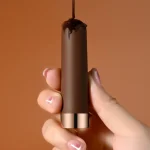 Artistic close-up of the chocolate-colored Mily intimacy tool being held, emphasizing its sleek aesthetic and luxury rose gold metallic base.