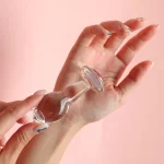 Clear glass anal plug held in hand against pink background Discreet Sex Toys Brand - HooHss